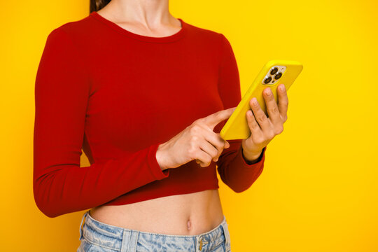 Fototapeta Young woman in red crop top uses yellow smartphone against bright yellow background showcasing casual fashion lifestyle and shopping promotion