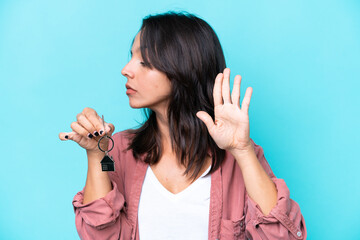 Young hispanic woman holding home keys isolated on blue background making stop gesture and...