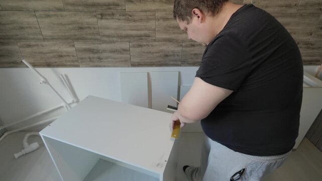 Skillful carpenter meticulously assembling a new white kitchen cabinet, carefully marking measurements with a pencil and square during a home renovation project and furniture installation.