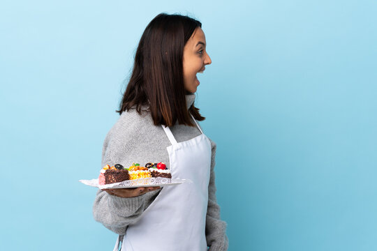 Pastry chef holding a big cake over isolated blue background laughing in lateral position.