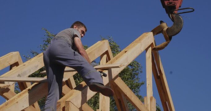 A young construction worker in overalls uses a hammer to install a wooden rafter system using a construction crane. The workers are erecting the roof frame for a new home under a clear blue sky.