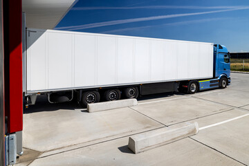 Blue truck with a white trailer parked in the yard of a logistics center. The truck is waiting to be loaded or unloaded into a warehouse. The photo was taken on a sunny day.
