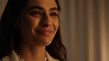 Close-up of a smiling woman with dark hair, wearing a light-colored jacket, looking towards the right.