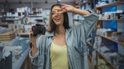 Young hispanic woman holding a dslr camera toward viewer with her hand and flashing a peace sign in a blue building; playful confidence.
