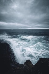 Fototapeta premium Dramatic ocean waves crash against rugged dark cliffs under a moody stormy sky, capturing nature's raw power and untamed beauty.