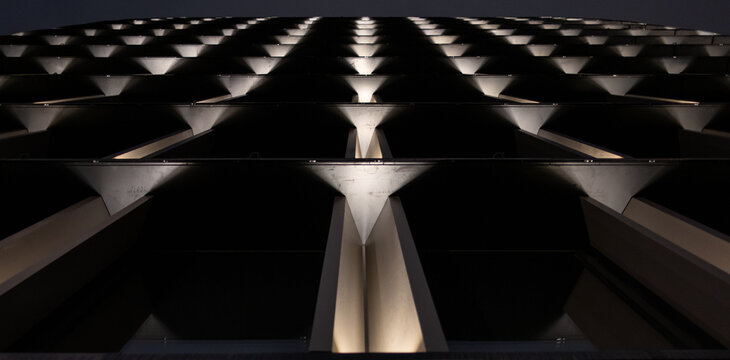 Low angle view of a modern building facade at night. The symmetrical, illuminated patterns create a futuristic and abstract architectural background with a strong sense of perspective.