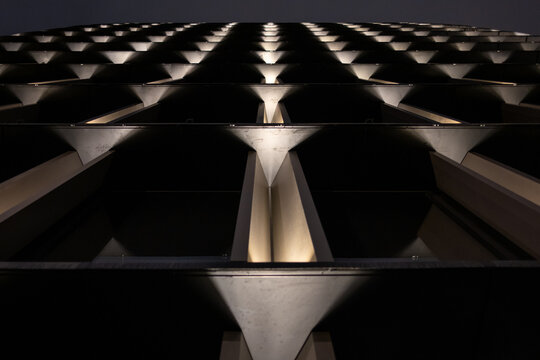 Low angle view of a modern building facade at night. The symmetrical, illuminated patterns create a futuristic and abstract architectural background with a strong sense of perspective.