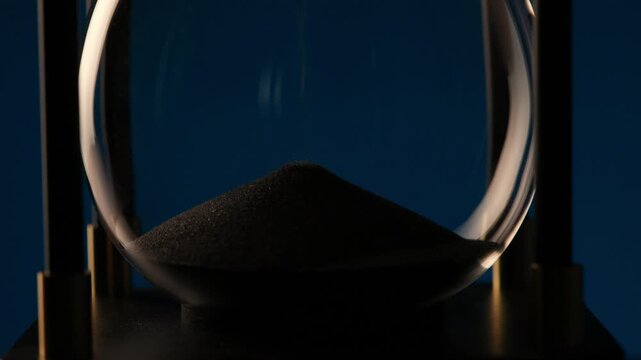 macro shot showing dark sand accumulating at the bottom of an hourglass, representing time running out and fleeting moments
