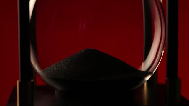 close-up of black sand piling up inside an hourglass on a red background, symbolizing the passage of time, patience, and inevitable change