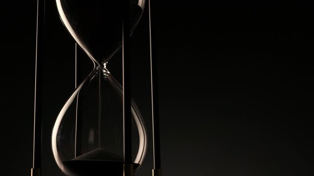 close-up video of dark sand flowing through a glass hourglass, capturing the silent and hypnotic passage of time in elegant motion.