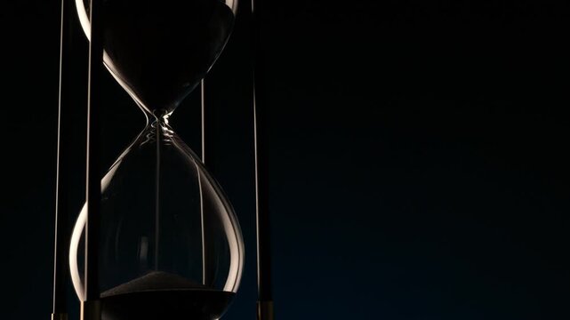 detailed video of dark sand falling in a transparent hourglass, representing the endless flow of time and fragile balance of life.