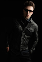 Obraz premium cool fashion man in black leather jacket with sunglasses holding hands in pockets