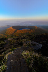Sunrise view from a mountain trail overlooking a volcanic crater lake surrounded by autumn colors and distant landscapes under a clear blue sky © Nuoh