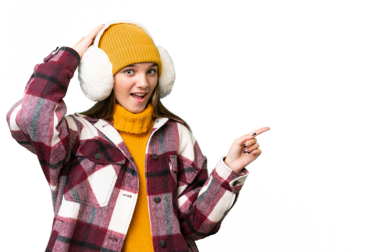 Teenager caucasian girl wearing winter muffs over isolated background surprised and pointing finger to the side