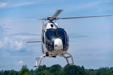 A white Airbus H120 helicopter hovers front-on above the runway, skids down and rotor blades spinning, framed by green treetops and a layered sky filled with altostratus and cumulus clouds. © Paul