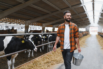 Farmer walking through barn while holding metal bucket and looking at cows © Serhii