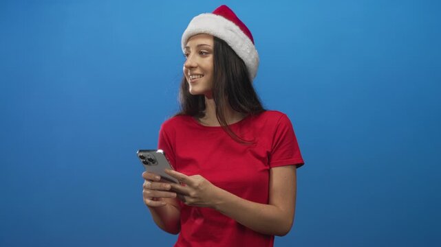 Woman wearing santa hat and red shirt holding smartphone and scrolling with a smile in blue studio; holiday cheer.