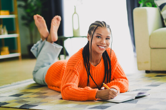Happy young woman lies on a rug at home writing in a notebook wearing a bright orange sweater in a cozy living room setting