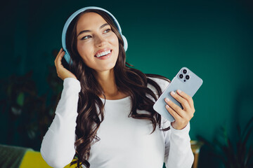 Happy young woman with headphones at home smiling while using phone in a modern living room setting...