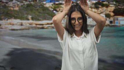 Woman with hands raised to head making antlers gesture on street beside beach and sea, wearing glasses and white shirt, smiling toward camera; playful.