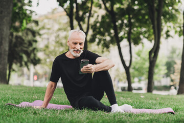 Older man with beard sits on a pink mat in the park outdoors using a smartphone during a morning workout in the city for wellness and fitness lifestyle