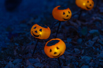 Line of glowing jack-o'-lanterns mounted on metal stands among fallen leaves at dusk, warm orange light against a deep blue evening backdrop.