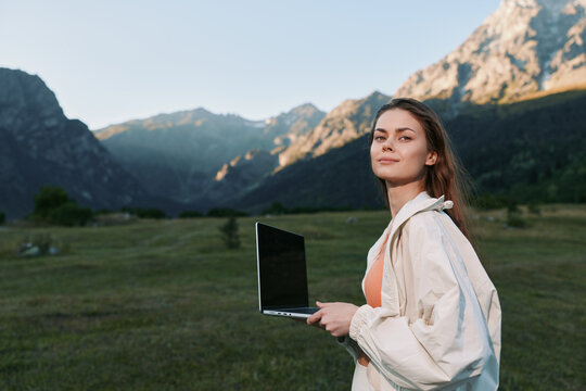 A composed outdoor professional holds a laptop in a green valley beneath towering peaks. She exudes confidence and focus, blending work and nature in a calm, productive moment.
