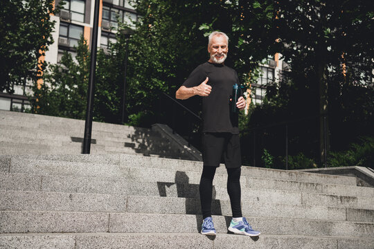 Older bearded man in black athletic outfit on outdoor city stairs giving thumbs up holding water bottle conveys fitness motivation and healthy lifestyle in urban park - Powered by Adobe