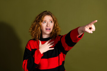 Young woman with red curly hair points forward while looking surprised against an olive green...