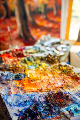 Close-up of a messy painter palette with thick dabs of oil paint in various bright colors. In the background, a blurry painting is visible, evoking creativity and inspiration.