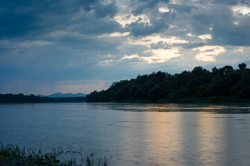 Rainfall over river during dusk, forest on riverbank and mountain in distance