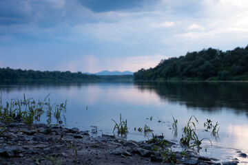 Landscape with muddy shore with some stones and grass, in background blurry forest on river bank and mountain in distance during rainfall at dusk