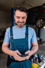 Mechanic checking messages on smartphone in garage with car repair equipment nearby