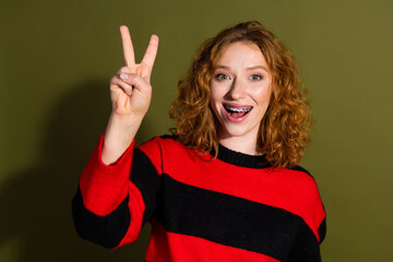 Young girl with red hair smiles and flashes peace sign wearing a red and black striped sweater against a khaki background