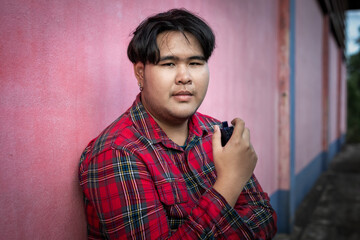 A young Asian man in a plaid shirt is standing alone smoking an e-cigarette and looking at the camera.