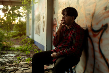 Young Asian man in a plaid shirt sitting and smokes a cigarette in abandoned building alone.