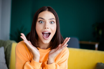 Fototapeta premium Joyful woman in orange sweater surprised expression at home in bright living room enjoying casual moment