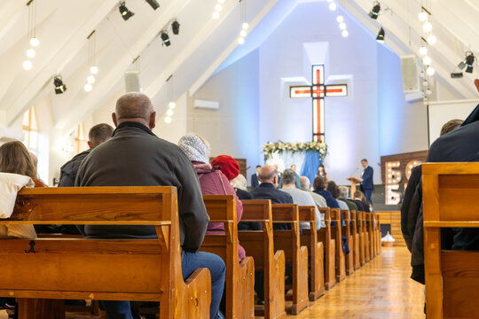 Congregation of diverse individuals seated in wooden pews, attentively listening to a speaker in a bright church interior, with a prominent cross illuminated in the background