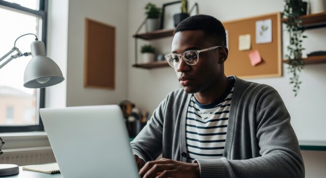 Focused African male student with glasses using a laptop for online education. Young professional working remotely from a home office. Digital business and freelance career development - Powered by Adobe