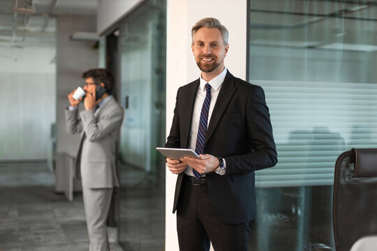 Smiling handsome businessman manager in suit standing in office with tablet computer. Colleague on the background