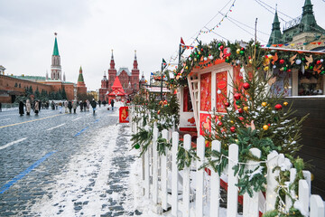 Festive Christmas market with decorated stalls and people walking in a snowy Red Square, Moscow. Winter holiday outdoor event for souvenir and gift shopping.