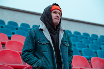 A man stands alone in a nearly empty stadium wearing a denim jacket and an orange beanie He appears deep in thought while looking around the cold colorful seats © Anton Dios