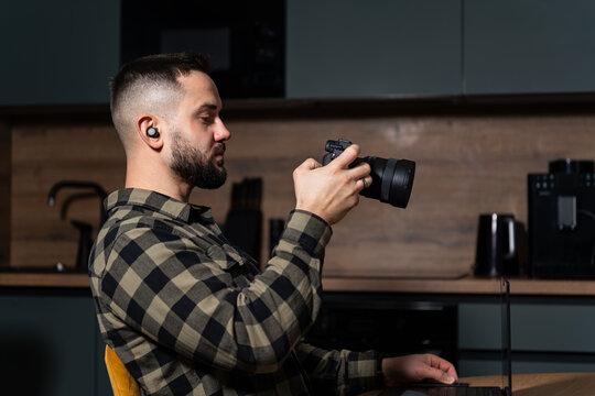 Bearded male photographer or content creator with earbuds holding a mirrorless camera, working on a laptop in a modern home office.