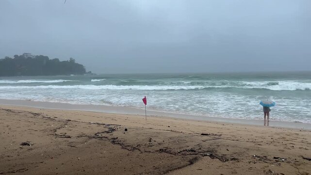 A person with an umbrella stands on a stormy beach during heavy rain. Rough ocean waves crash on the shore with a red warning flag indicating danger. Tropical monsoon weather concept