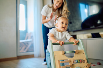 Happy mother cheering for her baby girl learning to walk using a wooden walker at home, celebrating development and growth