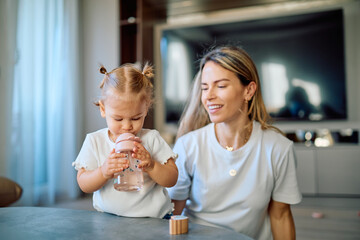 Adorable toddler girl drinking water from a sippy bottle at home, her mother smiling while watching. Family hydration