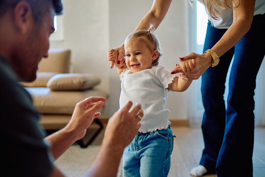 Smiling baby girl holding parents' hands, taking first steps indoors. Family bonding during a childhood development milestone