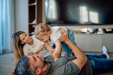 Obraz premium Father lifting laughing baby daughter while mother watches, enjoying fun family time together in their modern living room