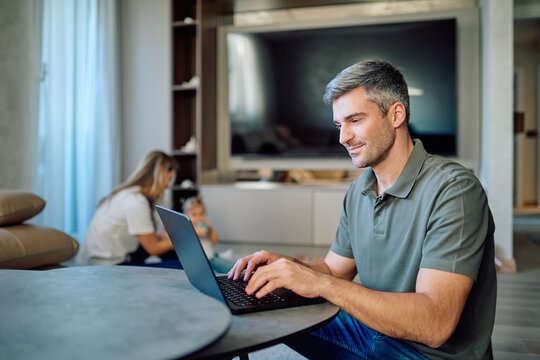 Man working on laptop at home, blurred family in background. Remote work balancing career and family life