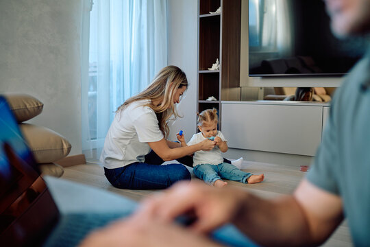 Busy parents balancing remote work and family life, with a mother assisting her toddler playing on the living room floor